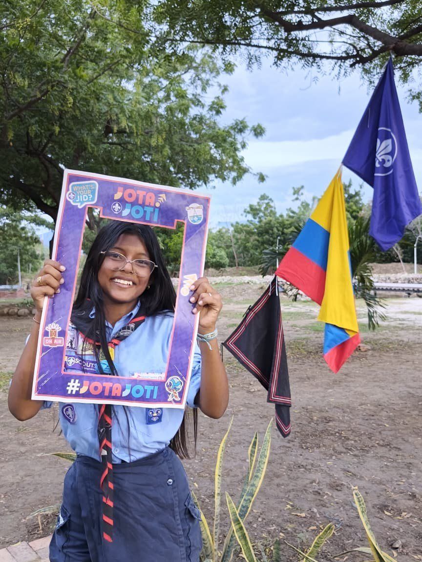 Una scout sonriente en el marco JOTA-JOTI, con banderas de Colombia y Scout